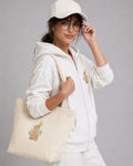 Person wearing a white hoodie and cap, holding a beige tote bag with a design on a gray background