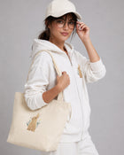 Person wearing a white hoodie and cap, holding a beige tote bag with a design on a gray background