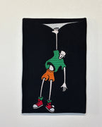 Embroidered patch of a skeleton in a green shirt and orange shorts hanging from a noose on a black background.