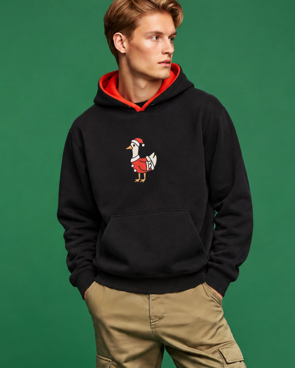 A man modeling a black pullover hoodie with a red hood lining and a silly santa goose embroidery on the chest, paired with tan cargo pants and white sneakers, set against a solid green studio background.