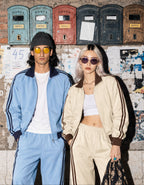 Two models posing in front of a brick wall with vintage mailboxes. The model on the left wears a light blue tracksuit with dark navy stripes and a black beanie.
